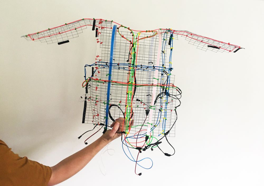 A photograph of a sculpture made out of multi-coloured wires in the shape of a sleeveless shirt being held up by a person's arm. 