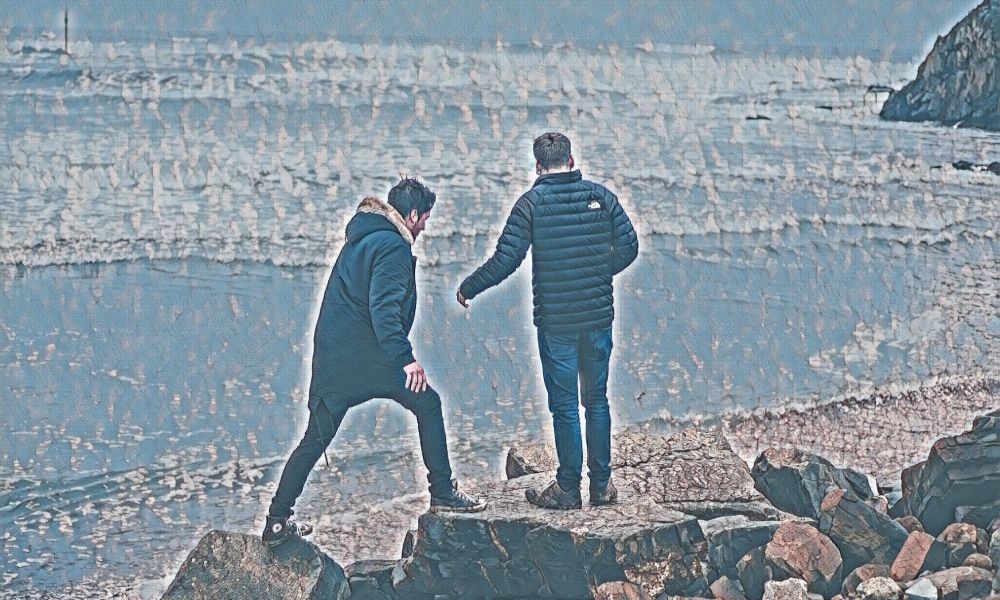 Two men dressed in winter jackets walking across rocks on a beach close to the water on a sunny day