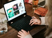 photograph of a woman typing on her black laptop situated on a wooden desk with a glass with ice and lime next to her