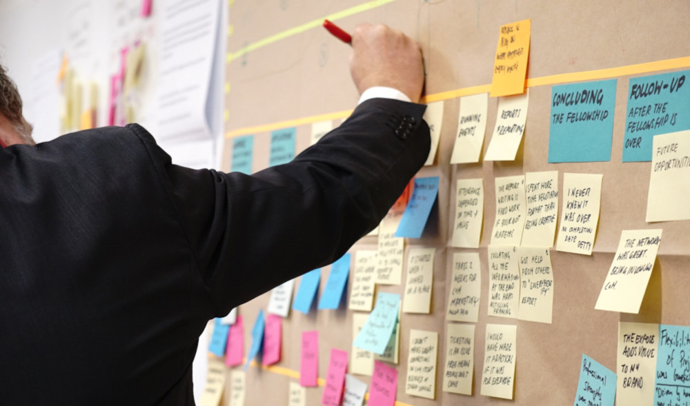 Many colourful sticky notes with writing on them on a cork board. A person is stretching out their hand to write on the board.