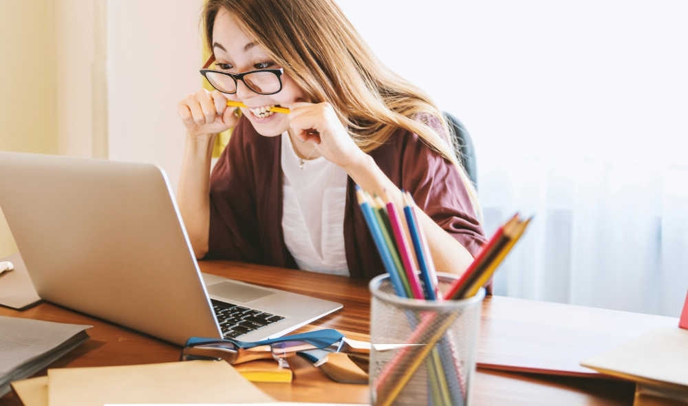 woman biting pencil while sitting on chair in front of computer during daytime