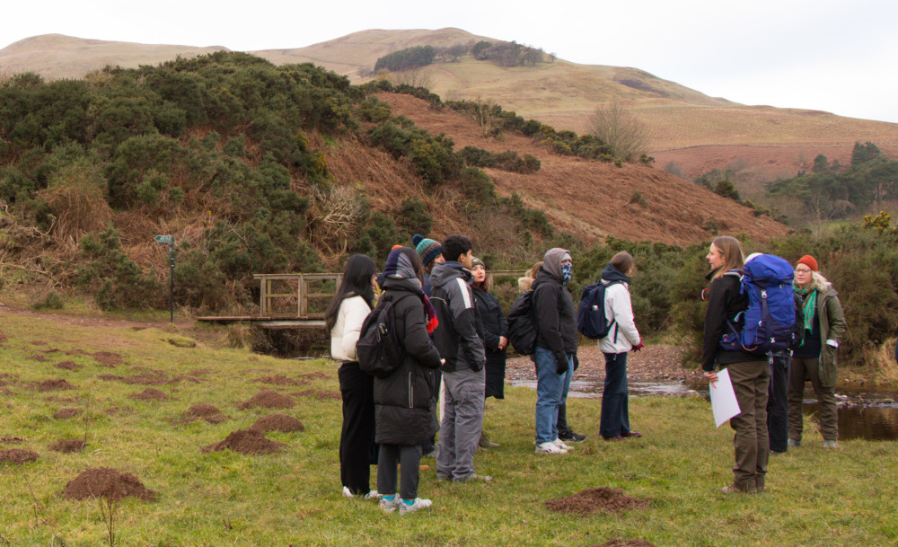 Students gathering on a hillside by a stream and bridge