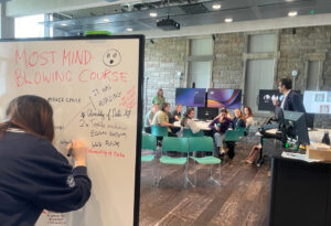 Student writing notes on a white board about the 'most mind blowing course'. Students sitting in chairs chatting behind her.