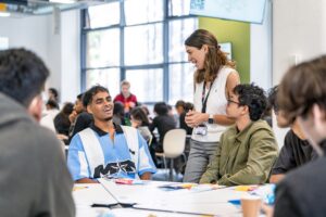 students chatting to a tutor around a table in a full classroom