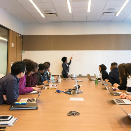 Students sitting around desk with teacher at front on whiteboard.