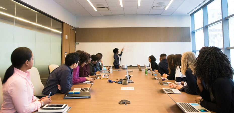 Students sitting around desk with teacher at front on whiteboard.