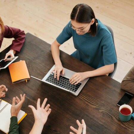 Student sitting around a desk, talking and typing on a laptop.