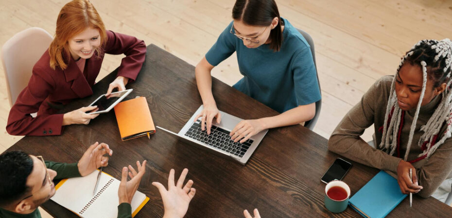 Student sitting around a desk, talking and typing on a laptop.