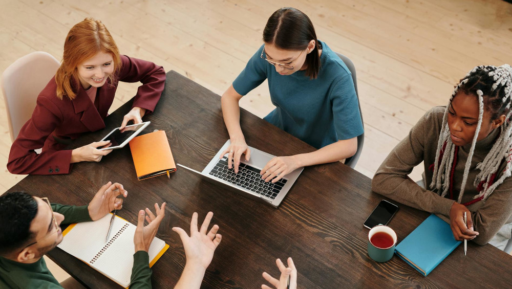 Student sitting around a desk, talking and typing on a laptop.