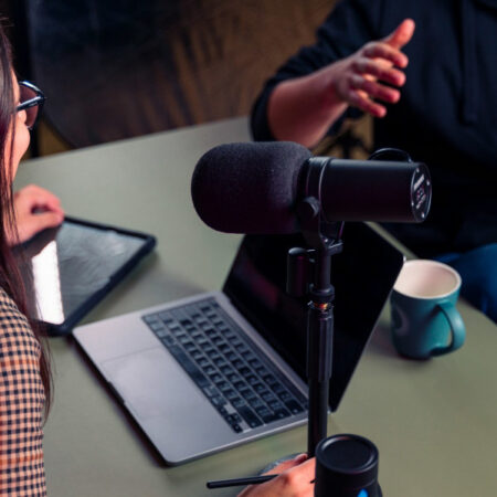 Photo of two people talking around a laptop and microphone to record a podcast