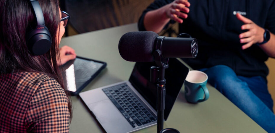 Photo of two people talking around a laptop and microphone to record a podcast