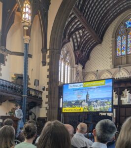 A man in a grey suit stands at a wooden podium in front of a large screen with the words 'Technician Commitment' and a picture of Glasgow University. There is a crowd of people sitting with their backs to the camera, watching him. The room they are in is large and old, with stained glass window and wooden balconies and decoration, similar to a church.