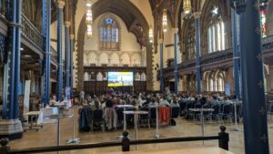 The image shows a large, grand room with an archway and a stained glass window at one end, with balconies and pillars along either side. There is a large screen at the end and in the middle, chairs with people in them facing away from the camera, looking at the person presenting. Along the sides of the room are tables with people sitting at them.