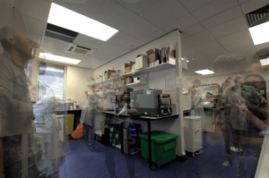 The image shows a laboratory with many shelves of equipment and boxes. Around the laboratory there are transparent images of many people, some wearing lab coats, some not.