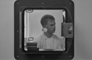 A black and white image of a person in a labcoat, viewed through a glass door. The person has short hair, and the background is a wall.