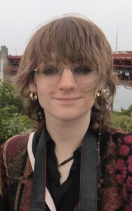 The image shows a person with short dark hair, round glasses, gold hoop earings staring at the camera. The person is Jaye Ross, the winner of the 2024 technician image competition. Jaye takes up the majority of the frame, with green bushes in the background and a small amount of the forth rail bridge in the top corner