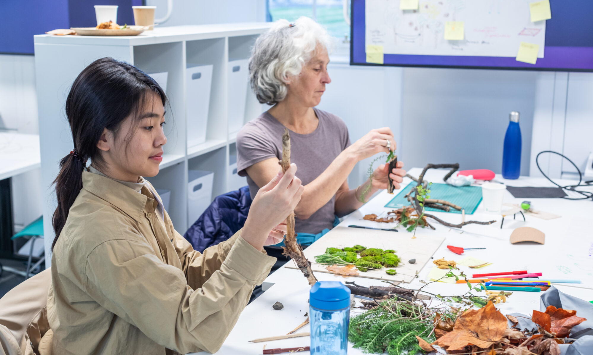 Two women making sculptures out of materials at a white table.