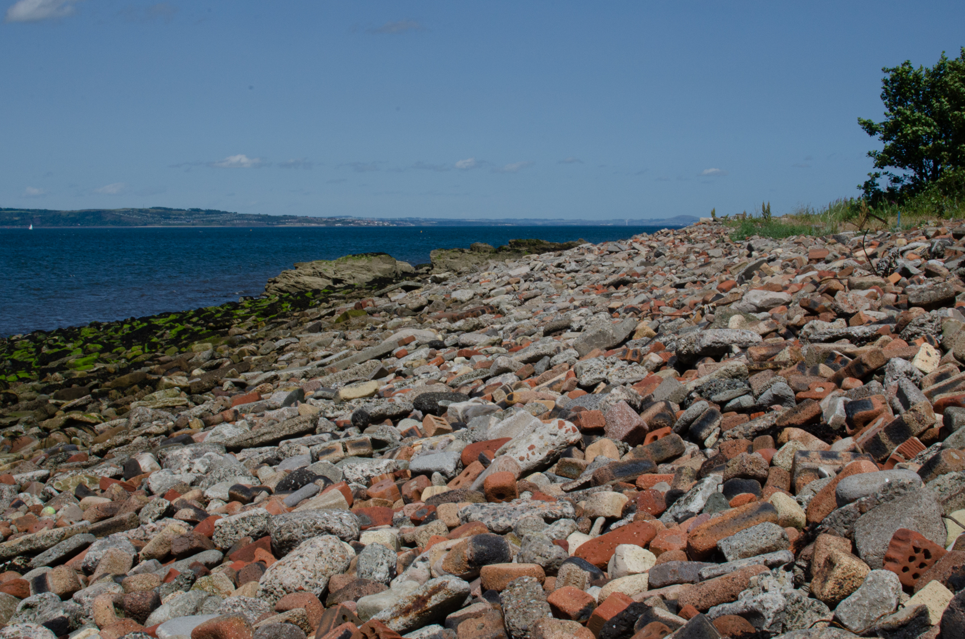 A view of Royston beach looking NE, towards the Firth of Forth. The fore and middle ground shows the slope of the beach with the sea at left, brick rubble covers everything. A large partially submerged rock formation can be seen in the distance and behind that, the Firth of Forth and a portion of Fife. The sky is blue and cloudless.