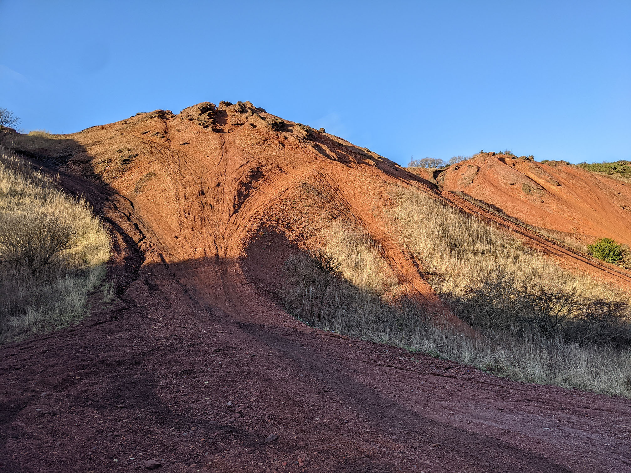 A view of an oil shale bing, coloured rusty orange, with a bright blue sky behind. The oil shle bings rises to a steep mound and is rutted with tracks and defiles. The foregound is in deep shadow.