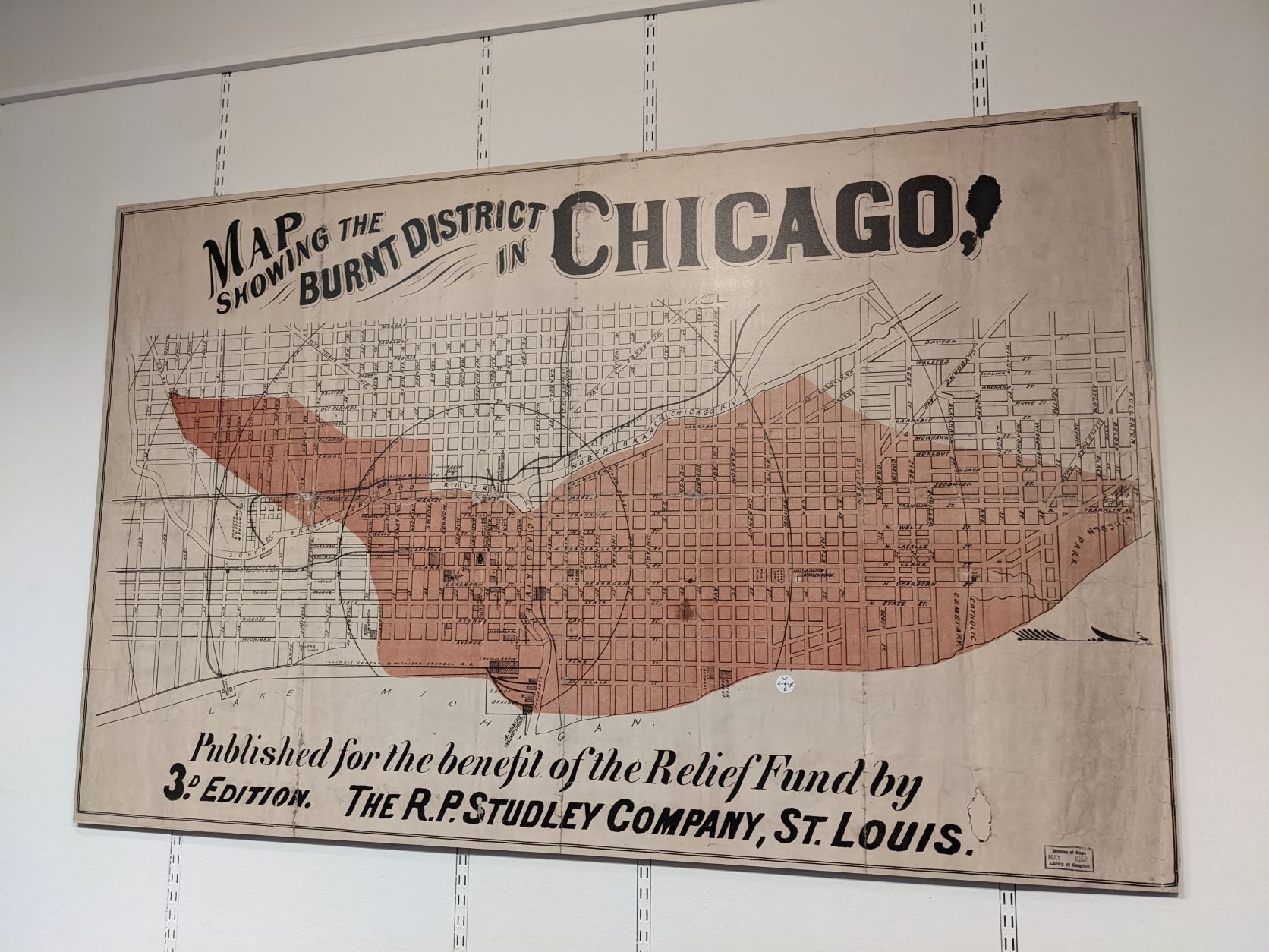 A map showing the burnt area of the fire of Chicago. The schematic grid map is tinged red with the burnt area shown and the centre point of the fire surrounded by regular concentric rings. The north of the map is to the right. The original printed caption reads: 'Map showing the burnt distric in Chicago, [beneath the map the caption then reads:] 'Published for the Relief Fund by The R.P. Studley Company, St. Louis. 3rd Edition.