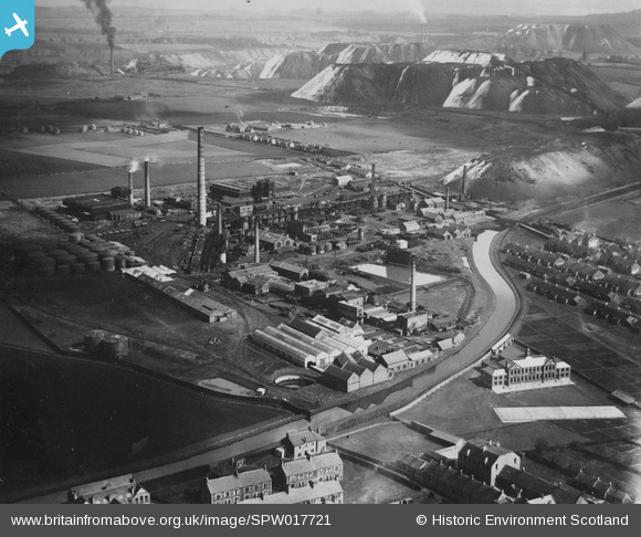 An oblique aerial photograph shows a complex of factories and chimneys dominating the lower half of the black and white image. A canal snakes across the bottom right hand corner of the image. A range of several angular spoil heaps (bings) loom in the background. An oilworks' chimney smokes at the top left of the image in the distance. Betweent he factories adn the mounds are a patchwork of agricultural fields.