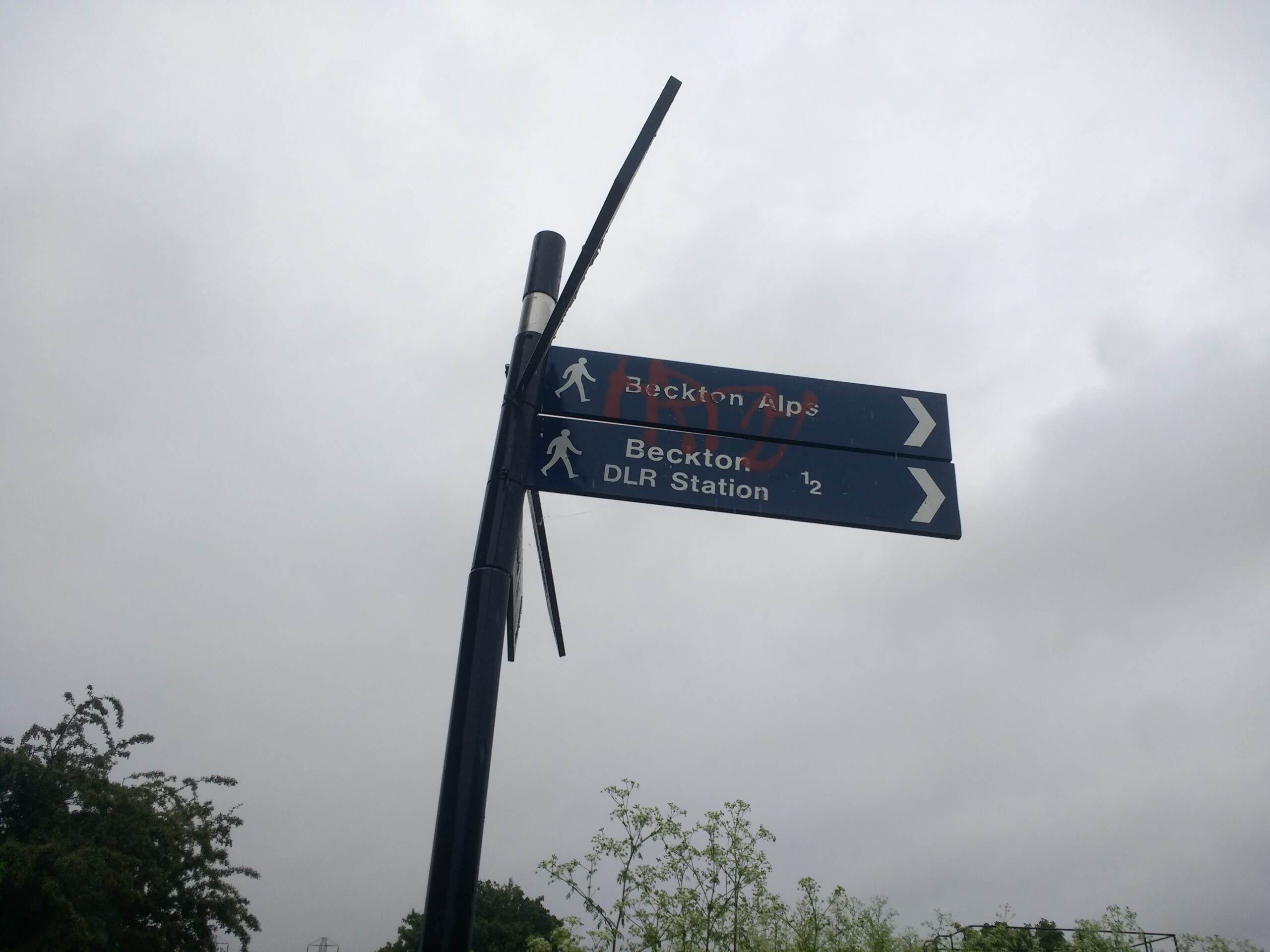 A photogrpah of the top section of a multi-directional signpost points right towards the 'BEckton Alp' and below, 'Beckton DLR Station'. The other signs pointing in the other directions are not visible.