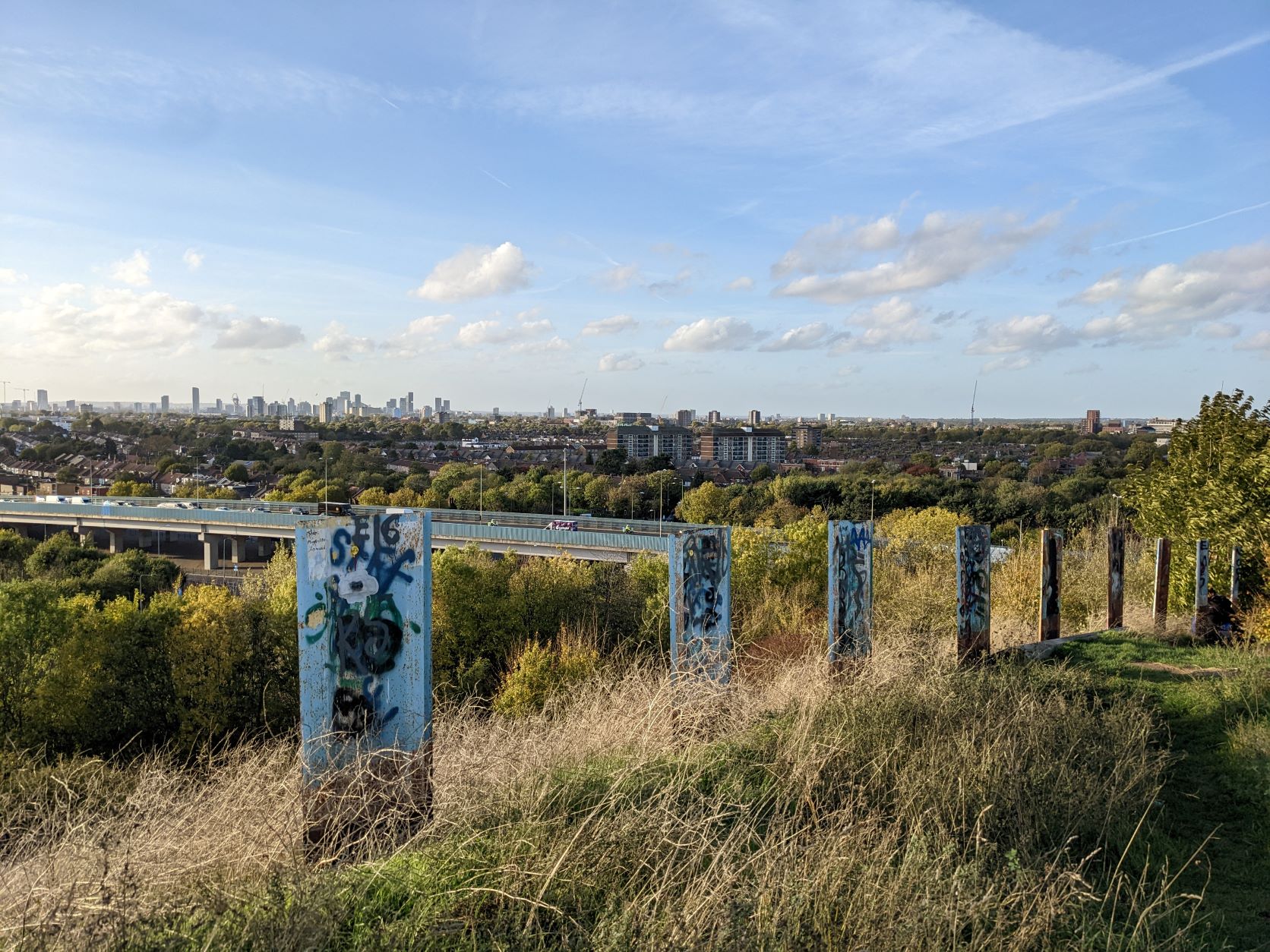 View from the top of the Beckton Alp towards Stratford, East London. The view takes in nine graffitied sheet piles standing in a row around 1m high emerging from the summit covered in long grass. The A13 motorway can be seen with the distant towers of Stratford on the horizon against a blue sky and evening light.