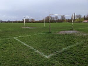 A view of several sets of football goal posts on the green fields of Hackney Marsh. The corner of the penalty box is seen painte dont he grass. The enarest goal mouth is muddy and worn and the sky is grey. A line of tres is in the background.