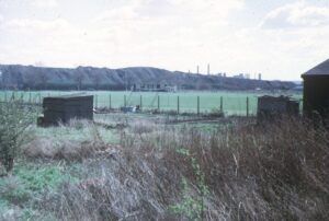 A large ridge of grey material about 25 m high runs across the centre of the image in front of a foreground of green - a football pitch. The immediate foreground has a small hut at left and two others at right. Distant gas works chimneys can be seen behind the ridge against a cloudy sky.
