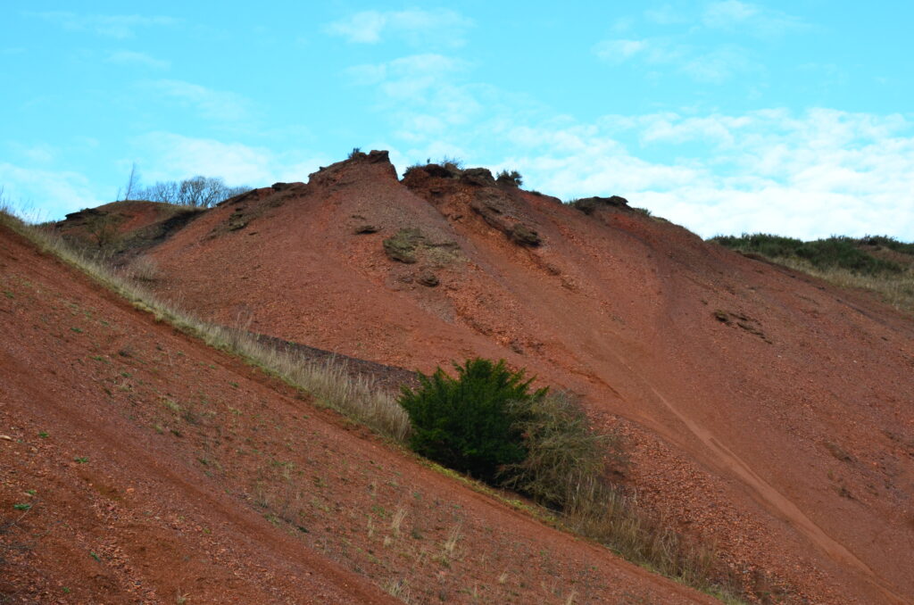 a mountain of shale waste blaes at greendykes bing. The shale blaes is red/orange and is piled in the majority of the frame into a blunt peak at centre, with a light blue sky above. A few small bushes pumctuate the otherwise seemingly barren hillside.