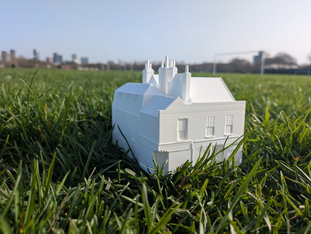 a small white plastic model house sitting on a grassy football pitch, tower blocks looming in the distance with a blue sky.