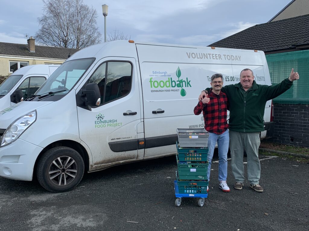 Two men hugging and smiling in front of an Edinburgh Food Project van, alongside a stack of donation crates from the Website & Communications team.