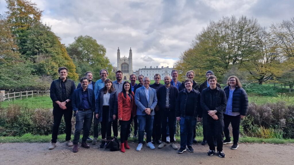 A mixed group of people standing with Cambridge University's King's College building at the back, trees on either side and a cloudy sky.