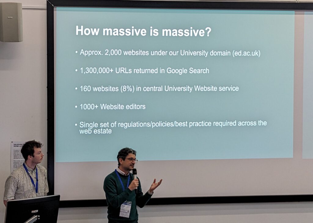 Stratos and Osh standing in front of a large screen listing numbers associated with the size of the University of Edinburgh Web Estate. Stratos is holding a microphone and talks with his left hand raised.