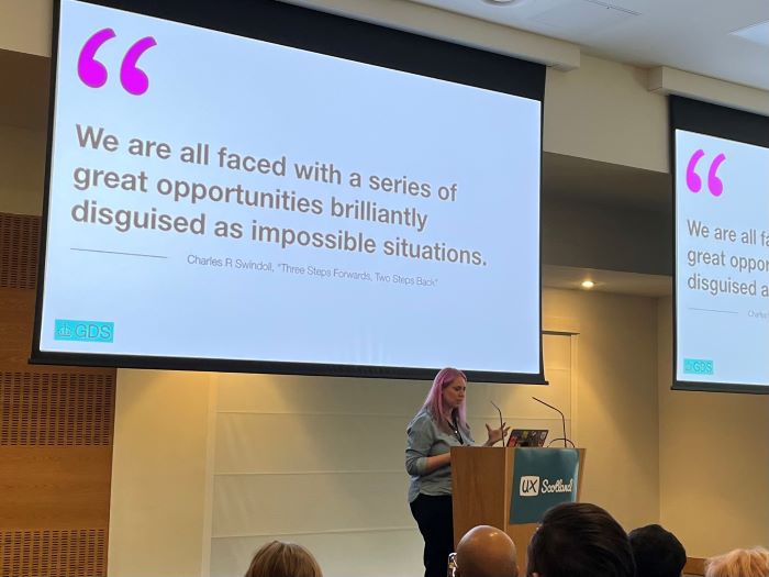 Laura presenting in front of a slide that says "We are all faced with a series of great opportunities brilliantly disguised as impossible situations." Quote is from Charles R Swindoll, Three steps forward, two steps back.