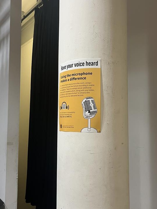A pillar in a lecture hall, with a yellow poster, titled 'Have your voice heard: Using the microphone makes a difference'. There is an icon of a microphone in the corner of the poster and a QR code that links to accessibility and lecture recording information.
