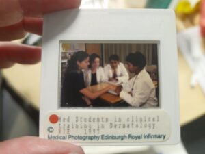 a photographic slides being held up white white edges with a small window, the photograph is a group of four male and females wearing white lab coats sitting around a table
