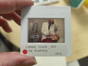 a photographic slides being held up white white edges with a small window, the photograph is a male in a white coat holding a handheld laser device to the face of a person lying down on a bed