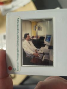 a photographic slides being held up white white edges with a small window, the photograph is a male in a white coat sitting at a computer