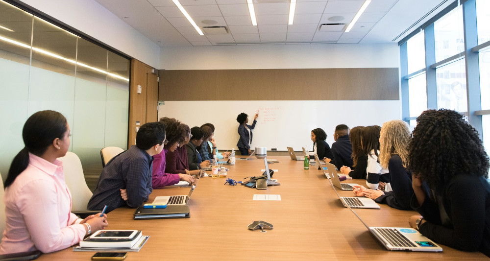 Students sitting around desk with teacher at front on whiteboard.
