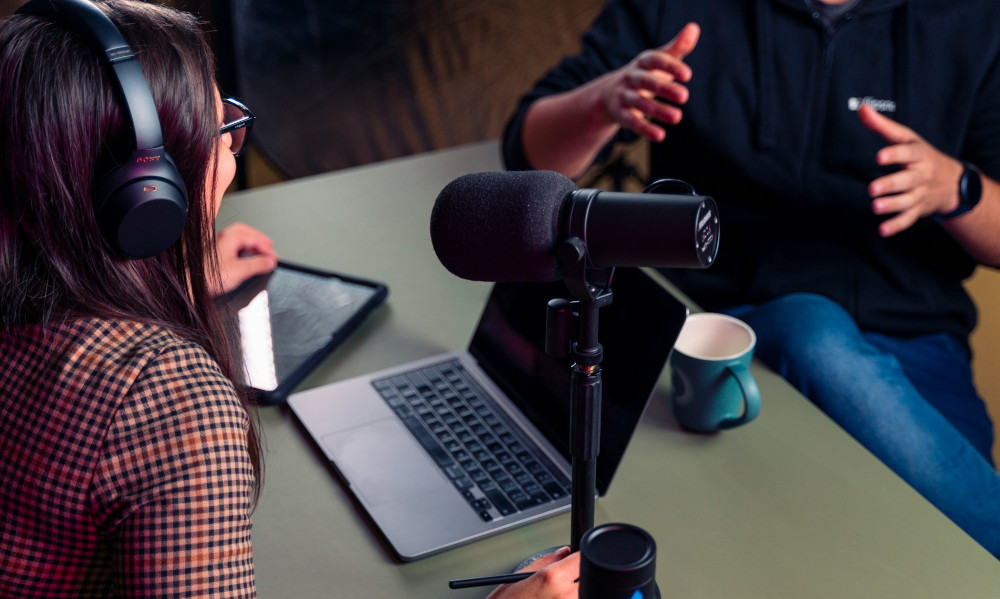 Photo of two people talking around a laptop and microphone to record a podcast