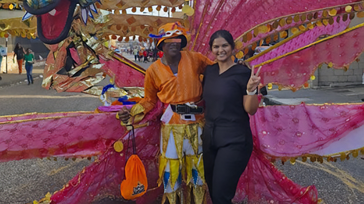 Arrianna Peters smiles and makes a peace sign beside a carnival performer in an elaborate orange and gold costume, with large decorative pink and gold carnival wings in the background.