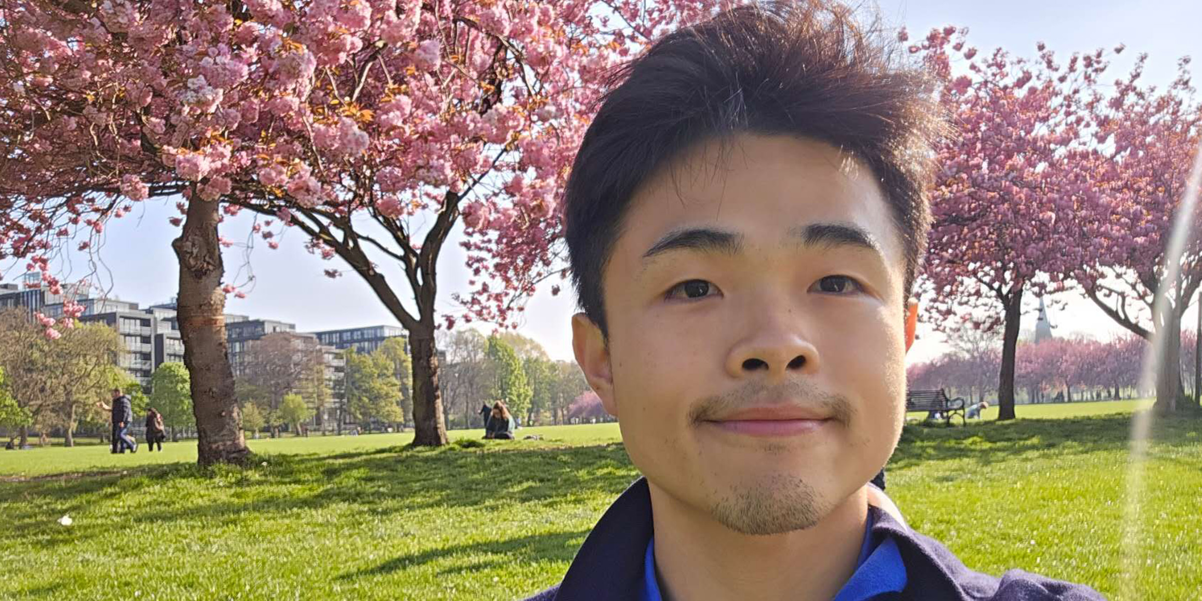 Student Ian standing in front of pink cherry blossom trees and smiling.